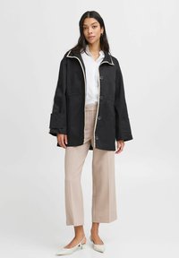 Zwarte waterdichte jas met witte details en grote knopen, met brede mouwen en een relaxte pasvorm, gecombineerd met beige broek en witte schoenen.