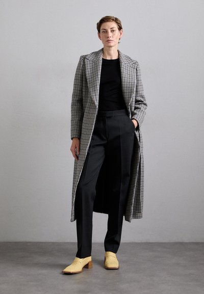 Woman wearing a long gray plaid coat, black top, black trousers, and yellow woven heeled shoes standing against a plain gray wall.