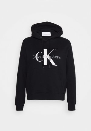 Čierna mikina s kapucňou, ktorá má klokanie vrecko, sťahovaciu kapucňu a výrazné biele logo "CK" s textom "Calvin Klein Jeans" pod ním. Mäkký materiál.