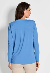 Blaues Langarmoberteil aus weichem Material mit Rundhalsausschnitt und Faltendetails auf der Rückseite. Das Design hat eine entspannte Passform und wird mit hellen Hosen kombiniert.