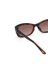 Occhiali da sole tartarugati con lenti marroni sfumate, angoli arrotondati e una sottile stanghetta nera con il logo "GUESS" impresso.