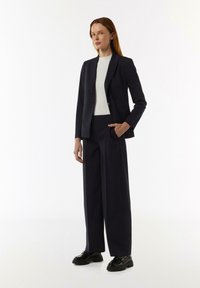 Navy gestreifter Anzug mit tailliertem Blazer und weiter Hose, kombiniert mit einem weißen Oberteil und glänzenden schwarzen Brogues.