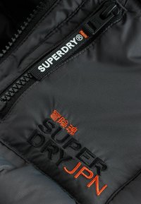 Sort polstret jakke i stof med en fremtrædende sort lynlås og broderede logoer i sort og orange, der læser "SUPERDRY" og "SUPERDRYJPN."