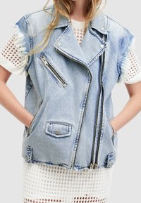 Ljusblå denim ärmlös väst med fransade kanter, två framfickor, sned dragkedja och silverfärgade detaljer. Lagd över en vit mesh-topp.