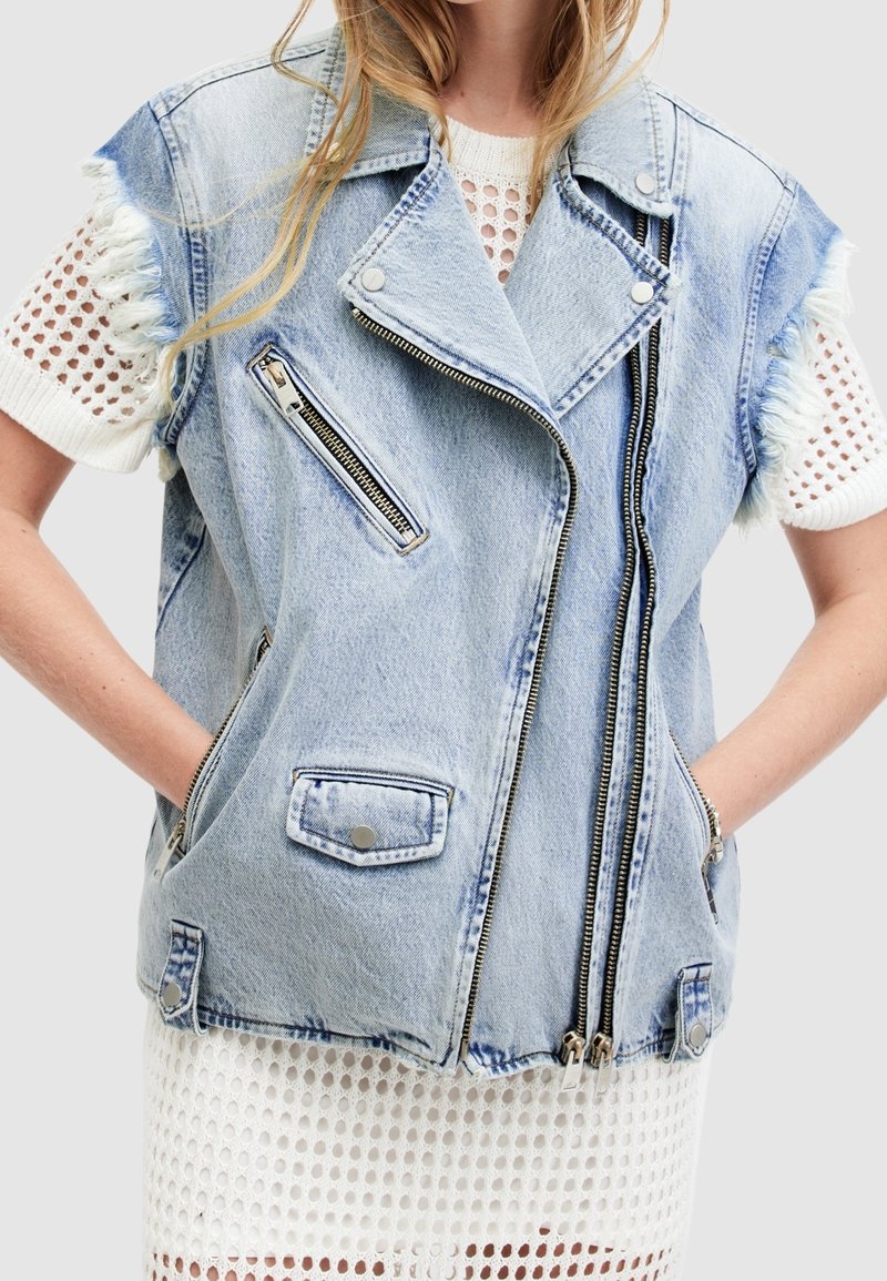 Ljusblå denim ärmlös väst med fransade kanter, två framfickor, sned dragkedja och silverfärgade detaljer. Lagd över en vit mesh-topp.