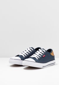 Granatowe trampki na płaskiej podeszwie z białym gumowym podbiciem i noskiem. Posiadają białe sznurówki, małą czerwoną metkę oraz brązowy akcent na pięcie.