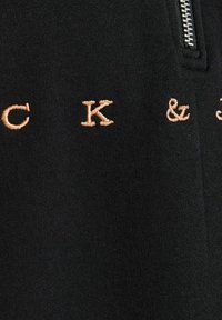 Tissu noir avec des lettres brodées beiges « C K & » et une fermeture éclair argentée visible sur le côté droit.