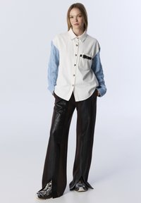Chemise boutonnée en tissu blanc avec manches tricotées en câble bleu clair, associée à un pantalon en cuir synthétique marron avec des fentes latérales.