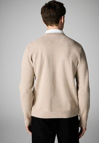 Beige strikket sweater med en tekstureret finish, med en rund halsudskæring og ribbede manchetter, parret med sorte bukser. Set fra ryggen.