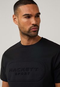 Schwarzes T-Shirt mit einem strukturierten, geprägten Logo "HACKETT SPORT", das zentriert auf der Brust sitzt, mit einem runden Ausschnitt und kurzen Ärmeln.