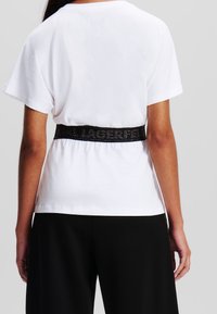 Bílé tričko s krátkým rukávem a černým elastickým páskem s výrazným potiskem. Měkká látka s volným střihem, kombinováno s černými kalhotami.