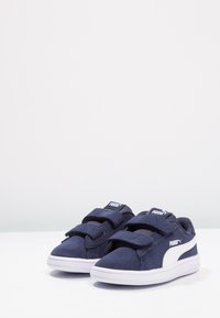 Zapatos de ante azul marino con acentos blancos y dos correas de velcro, con punta redonda y suela de goma blanca. El diseño incluye un logotipo.