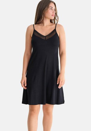 Femme portant une robe noire sans manches avec des bretelles fines et un panneau en V transparent à l'encolure, debout devant un fond uni.