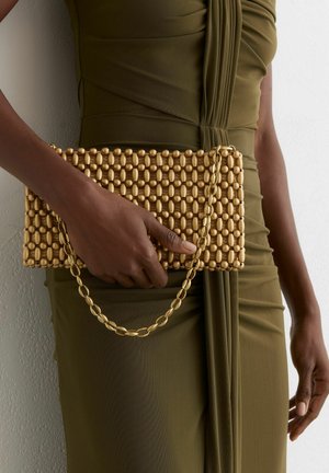 Oseba s temno poltjo v olivno zeleni naborkasti obleki drži pravokotno zlate perlice torbico z verižnim trakom pred ozadjem teksturirane stene.