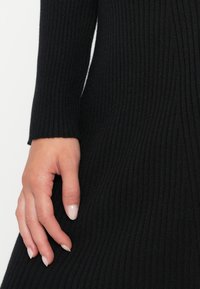 Main avec un vernis à ongles clair reposant sur un tissu noir côtelé en tricot à manches longues, montrant la manche et le côté d'un vêtement.