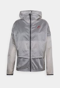 Lehká šedá bunda s kapucí, s černým mřížkovaným vzorem, zipovým zapínáním a červeným logem na levé hrudi. Obsahuje elastické manžety a lem.