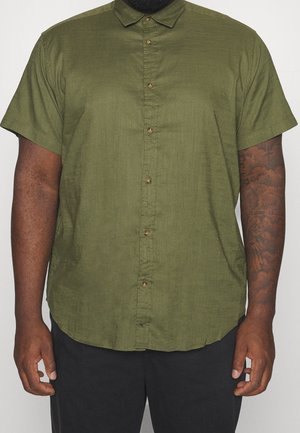 Mann trägt ein olivgrünes kurzärmeliges Hemd mit Knöpfen, hat Tätowierungen am rechten Unterarm und eine schwarze Hose, steht vor einem einfarbigen Hintergrund.