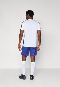 White athletic shirt with black side panels, blue shorts with red trim, white socks, and black soccer shoes. Model shown from behind.