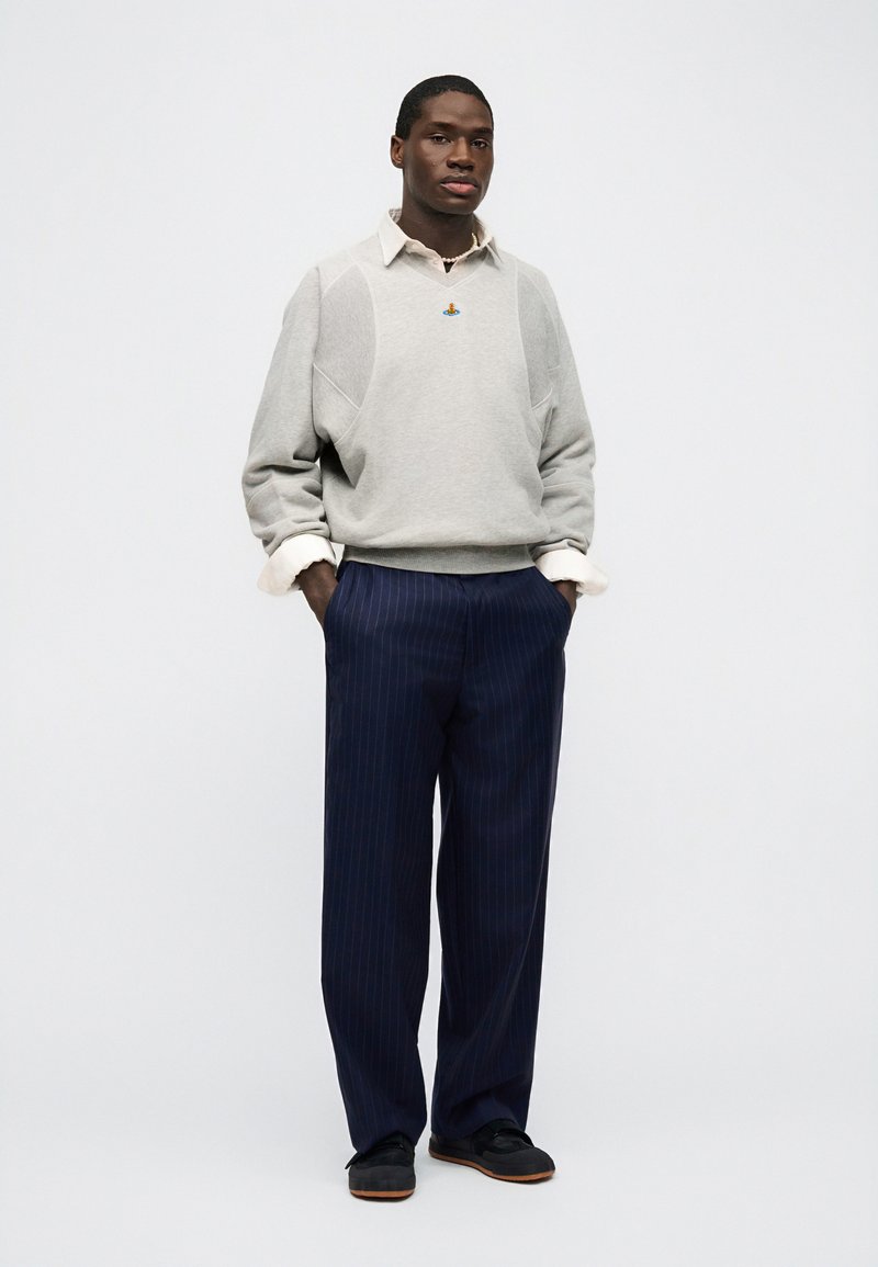 Uomo che indossa un maglione grigio chiaro sopra una camicia bianca con colletto, pantaloni blu navy a righe sottili e scarpe nere, in piedi con le mani nelle tasche.