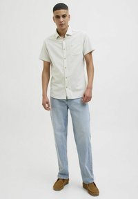 Chemise à manches courtes, de couleur claire, avec une poche, fermeture à boutons, motif subtil, assortie à un jean bleu clair et des chaussures à enfiler marron.