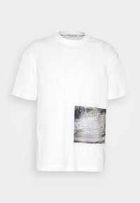 Bílé bavlněné tričko s krátkými rukávy a kulatým výstřihem, na kterém je obdélníková šedá grafika s nápisem "Calvin Klein Jeans."