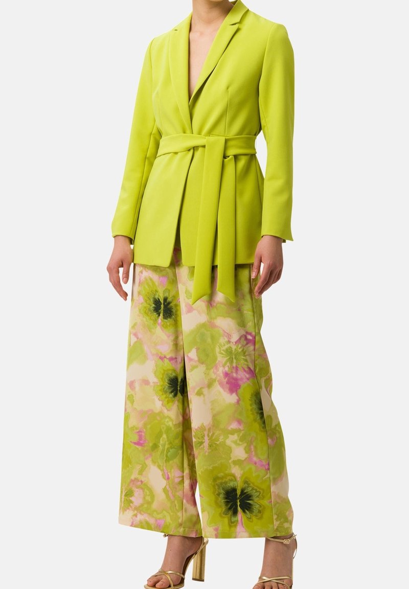 Femme portant un blazer ceinturé vert citron vif et un pantalon large à motifs floraux verts et roses, assortis à des sandales à talons hauts dorées.