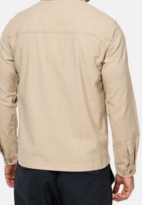 Homme portant une chemise beige à manches longues avec poignets boutonnés et un pantalon noir, vu de dos sur un fond uni.