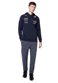 Granatowy hoodie z czarnymi i niebieskimi akcentami, z kieszenią kangurzą i logo. Połączony z szarymi spodniami i czarnymi butami sportowymi.