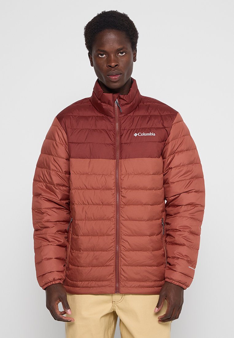 Chaqueta acolchada en marrón rojizo con un cuello alto y cremallera completa. Presenta un diseño bicolor y un logotipo visible de la marca en el pecho.