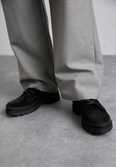 Chaussures en daim noires avec semelles en caoutchouc, présentant un design à lacets et des détails de couture subtils, assorties à un pantalon gris clair.
