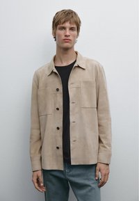 Light beige suede jacket with a collared neckline, two large front pockets, and button closures. Worn over a black shirt and denim pants.