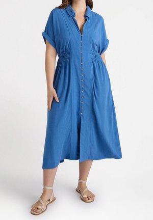 Kvinde iført en blå midikjole med knapper foran, med indsnøring ved taljen og opkørte korte ærmer, kombineret med beige flade sandaler med ankelrem.