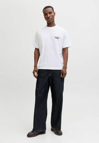 Λευκό βαμβακερό T-shirt με την ένδειξη "ORIGINALS" τυπωμένη στο στήθος, σε συνδυασμό με σκούρα μπλε φαρδιά τζιν παντελόνια και καφέ παπούτσια.