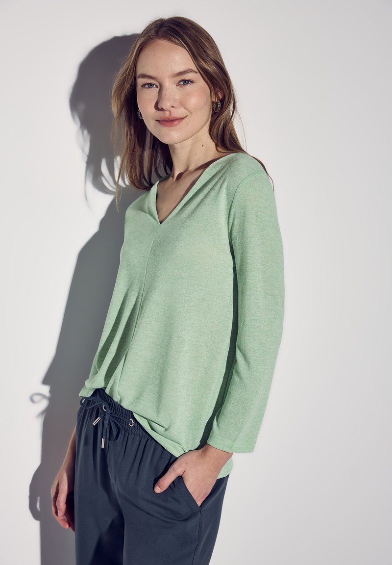 Top de color verde claro, de cuello en V, de manga larga y ajuste holgado. Combinado con pantalones oscuros con cordón. Textura suave sobre un fondo neutro.