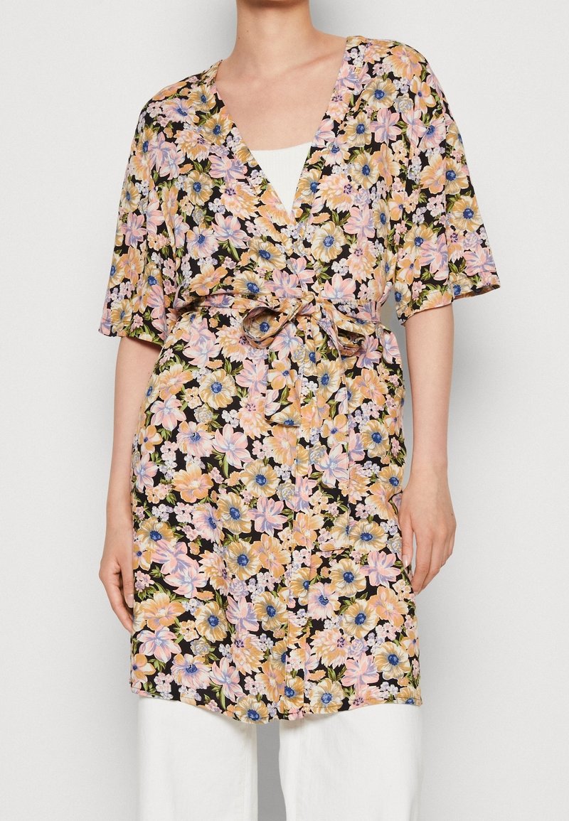 Femme portant une robe portefeuille fleurie à manches courtes avec un lien à la taille, par-dessus un top blanc et un pantalon blanc.