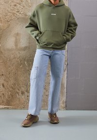 Felpa verde oliva con tasca frontale e logo bianco, abbinata a jeans a gamba ampia blu chiaro e scarpe da ginnastica marroni con accenti rossi.