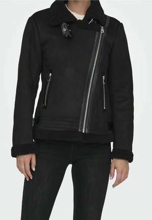Czarna kurtka ze sztucznego owczej skóry z asymetrycznym srebrnym suwakiem, kieszeniami na zamek, detalami z klamrami na kołnierzu i mankietach, noszona na białej bluzce i czarnych spodniach.