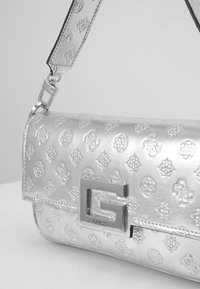 Silberne Handtasche aus Kunstleder mit geprägten Mustern, einem metallischen Logoschnappverschluss und einem abnehmbaren Tragegurt. Glatte Textur mit einem eleganten Design.