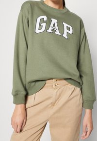 Felpa verde con logo "GAP" ricamato in bianco. Tessuto morbido, polsini e orlo a costine. Abbinata a pantaloni beige chiaro con chiusura a bottone.
