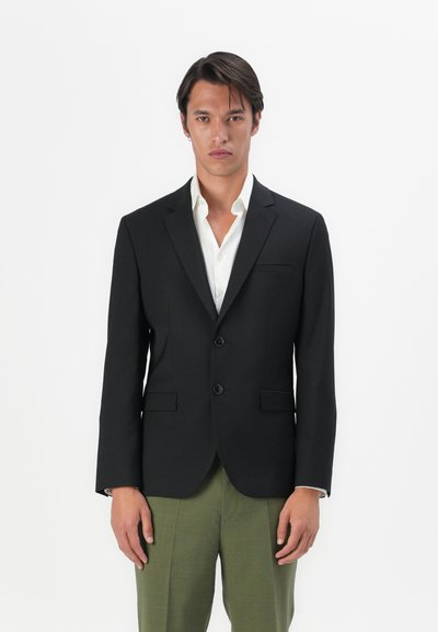 Blazer noir ajusté avec revers en pointe, deux boutons et une poche poitrine, porté sur une chemise blanche. Stylé avec un pantalon vert.