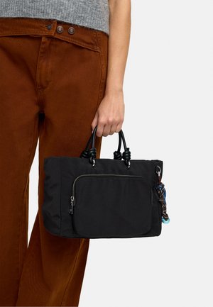 Ręka trzymająca czarną torebkę z przednią kieszenią na zamek i z supłowanymi uchwytami, noszona z rdzawymi spodniami i szarą bluzką na białym tle.