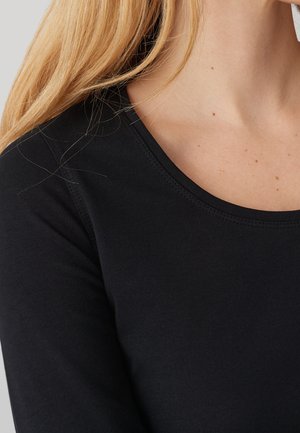 Blond osoba ubrana w dopasowaną czarną koszulkę z długim rękawem i zaokrąglonym dekoltem, odsłaniającą szyję i obszar ramion.