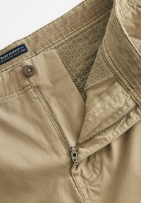 Pantaloni beige con zip e chiusura a bottone. All'interno presentano un rivestimento di tessuto a fantasia. Texture liscia con dettagli di cucitura robusti.