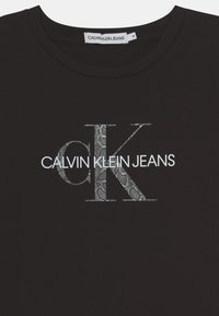 Czarna koszulka Calvin Klein Jeans z dużym logo "ck" w wzór wężowej skóry oraz białym napisem "CALVIN KLEIN JEANS" na klatce piersiowej.