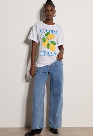 Frau trägt ein weißes Oversized-T-Shirt mit gelben Zitronen und dem Schriftzug „Capri Italy“, kombiniert mit lockeren hellblauen Jeans und schwarzen Stiefeln.