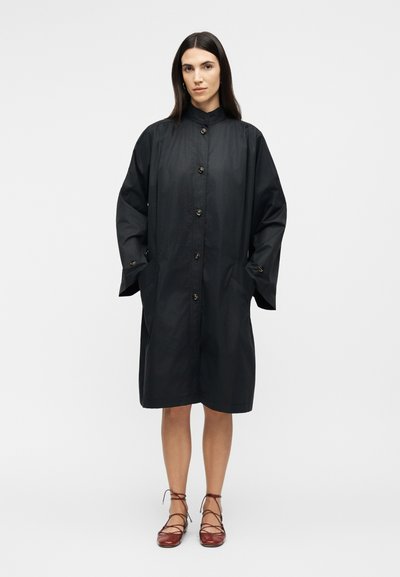 Femme portant un manteau noir ample à boutons, manches longues et poches, associé à des chaussures plates marron à lacets, debout devant un fond blanc.