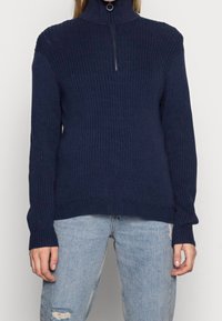 Pull en maille bleu marine avec un col montant à zip, texture côtelée et manches longues, associé à un jean bleu clair déchiré.