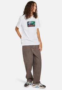 T-shirt bianco di cotone con stampa grafica di montagne e alberi in verde e nero, abbinato a pantaloni larghi marroni e sneakers multicolori.