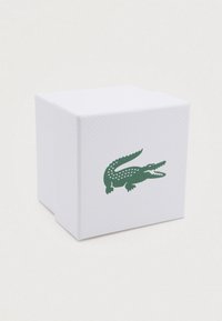 Boîte blanche avec une texture en relief, présentant un logo de crocodile vert sur un côté. Forme carrée, design épuré, apparence minimaliste.