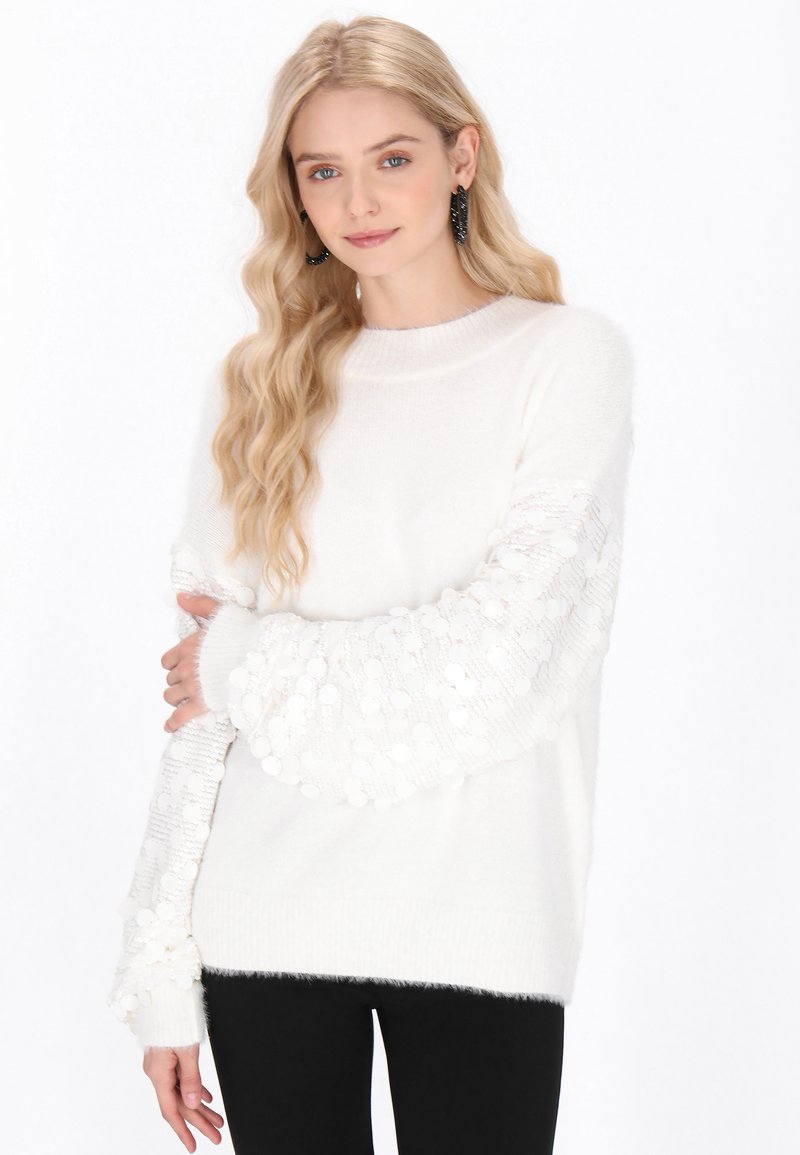 Weißer Pullover mit strukturierten, flauschigen Ärmeln, die kreisförmige Muster aufweisen. Gerippter Halsausschnitt und Bündchen, kombiniert mit schwarzen Hosen.